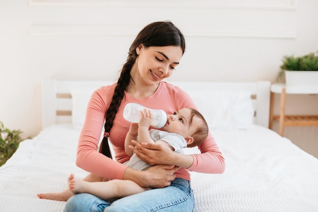 Cute infant baby drinking milk from bottle while lying on mothers hands woman sitting on bed at home