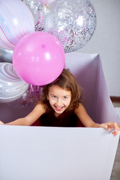 분홍색 드레스를 입은 귀엽고 즐거운 어린 소녀가 집 생일 파티 깃발에 풍선이 있는 큰 선물 상자 밖을 내다보고 있습니다. 생일 축하합니다. 축하
