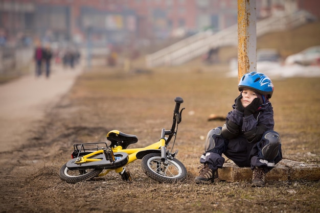 ヘルメットをかぶったかわいい子供の男の子が黄色の自転車を持って通りに座り、遠くを見ている