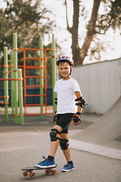 Cute kid boy in a helmet standing in a special area in skatepark and step on skateboard Child performs tricks Summer sport activity concept Happy childhood