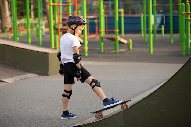 Cute kid boy in a helmet standing in a special area in skatepark and step on skateboard Child performs tricks Summer sport activity concept Happy childhood