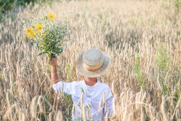 Ragazzo carino con cappello di paglia con margherite in mano che camminano sul campo di grano di segale