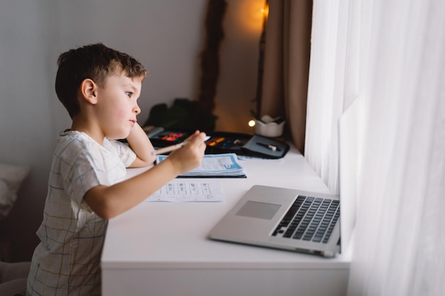 Cute kid boy studying at home with laptop and doing school homework