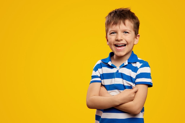 Cute kid boy wearing blue striped polo laughing with arms crossed against yellow background
