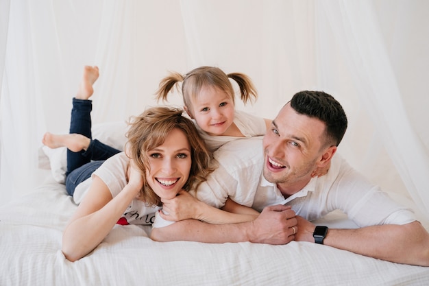 Cute kid daughter and dad tickling mom having fun good time playing together at home, happy parents and little child girl enjoying funny activity and communication, family laughing relaxing