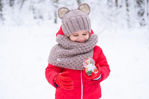 cute kid girl in pink snowsuit and knitted hat and scarf holds snow ball in winter forest