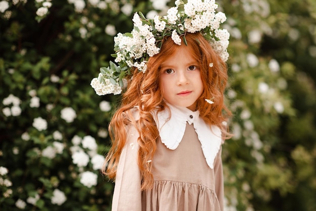 Cute kid girl with long curly red hair wear floral wreath and stylish dress over nature background