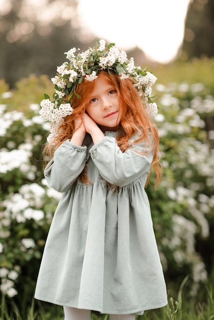 Cute kid girl with long curly red hair wear floral wreath and stylish dress over nature background