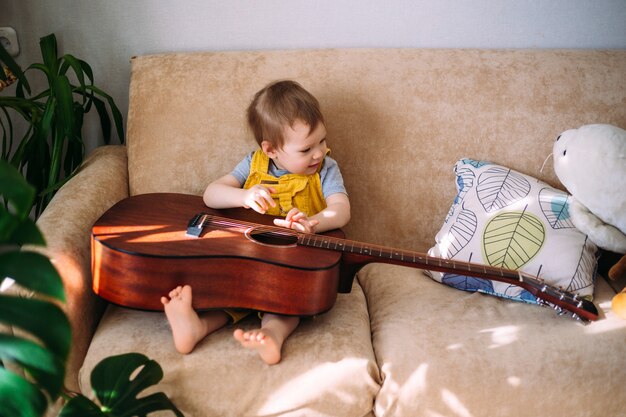A cute kid plays with a big acoustic guitar at home on the couch.