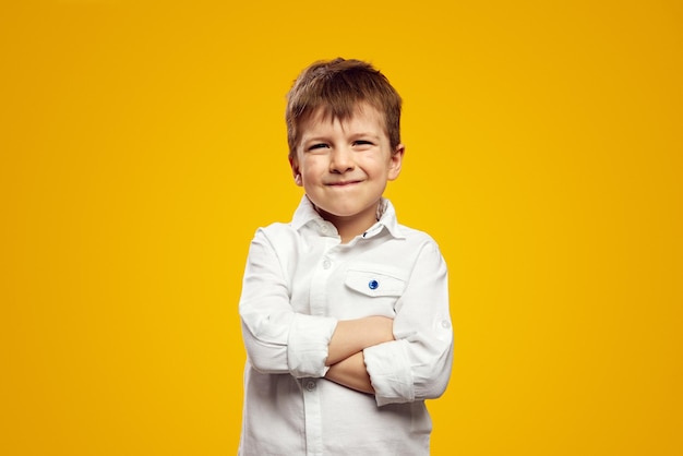 Cute kid wearing white shirt smiling for camera while keeping hands crossed against yellow backdrop