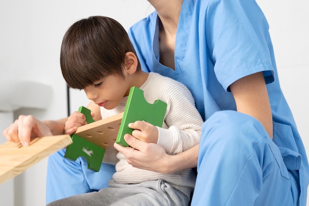 Cute kid with disability playing with developing toys while is being helped by physiotherapist in re...