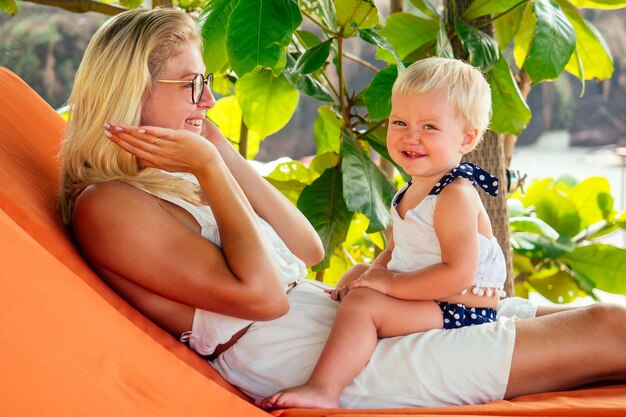 Симпатичный ребенок и молодая блондинка-родительница, играющая в прятки, шутит. Женщина и маленькая светловолосая девочка, годовалый ребенок на пляже на фоне тропических зеленых деревьев. День рождения матери
