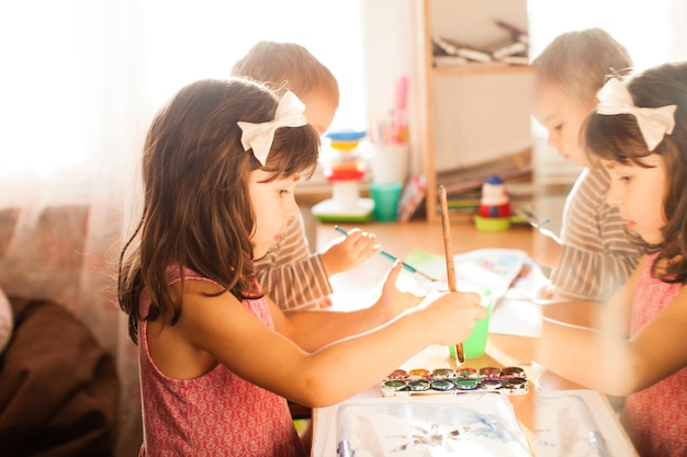 Bambini carini che dipingono con pennello e vernici colorate adorabile ragazza di 4 anni che disegna su carta bianca vicino alla finestra in una stanza luminosa