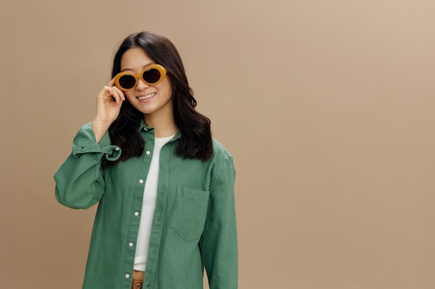 Cute korean smiling young woman in khaki green shirt stylish\
touches eyewear posing isolated on over beige pastel studio\
background cool fashion offer sunglasses ad concept