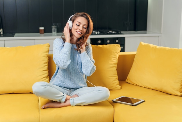 Signora carina con capelli castani ondulati in maglione blu che si siede sul comodo divano e godendo piacevoli melodie su auricolari nella cucina contemporanea.