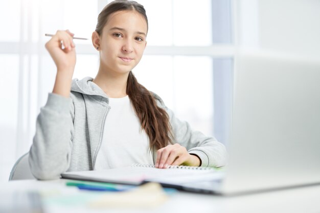 Cute latin teenage girl smiling at camera, using laptop while studying at home. Distance education, home schooling concept