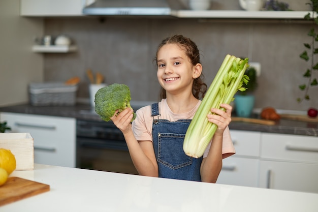 Cute laughing and curly little girl stand near table in the kitchen, holding greens vegetables in hands.