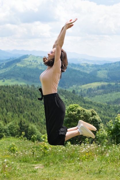 Cute laughing girl is jumping against the backdrop of mountains and forests. Sunny summer day