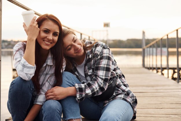 Photo cute lesbian couple sits together near the lake with cups of drink in hands and embracing each other