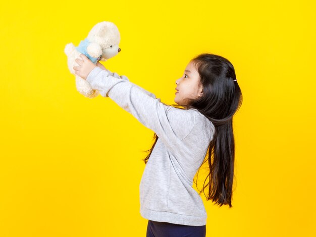 Cute liitlw Asian girl holding and playing with teddy bare doll on yellow background. Concept of kid funny and imagination.