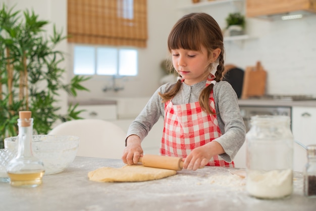 Милая маленькая 4-летняя девочка с косичками раскатывает тесто скалкой на кухонном столе.