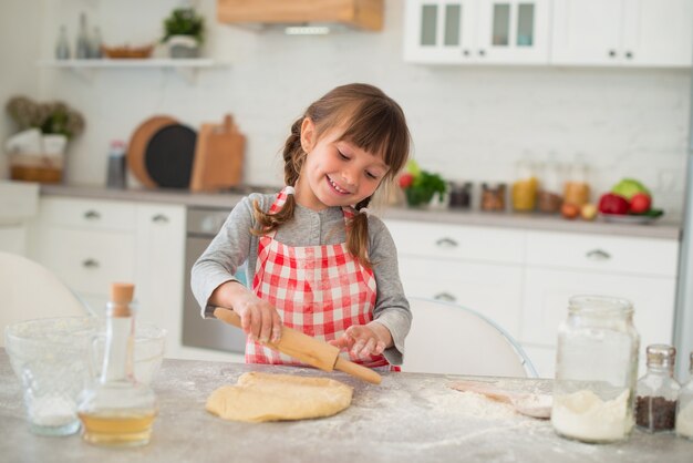 おさげ髪のかわいい4歳の女の子は、台所のテーブルに麺棒で生地を広げます。