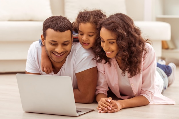 Cute little Afro-American girl and her beautiful young parents using a laptop and smiling while lying on the floor in the room.