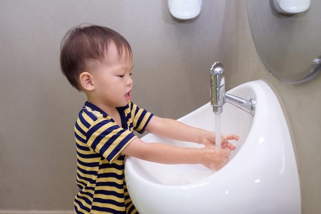 Cute little Asian 2 years old toddler baby boy child washing hands by himself on white sink and water drop from faucet in public toilet / bathroom for kids, Sanitation / hygiene concept