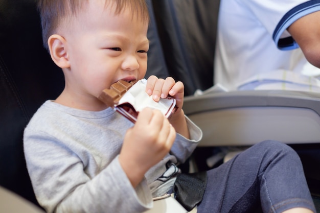 Cute little Asian 2 years old toddler boy child eating chocolate bar during flight on airplane. Flying with children concept