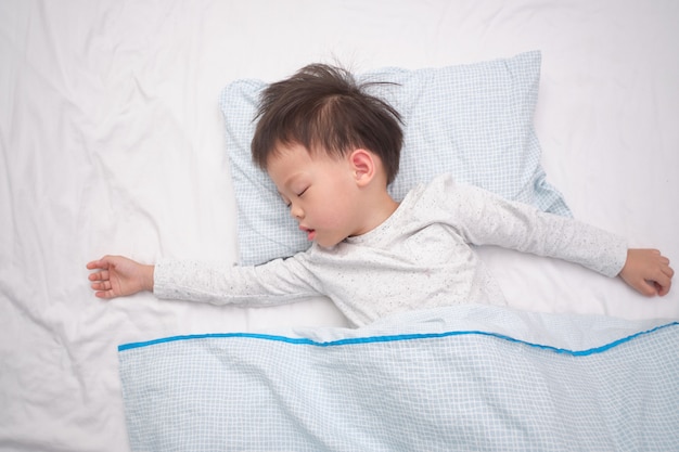 Cute little Asian 3 - 4 years old toddler boy kid in pajama taking a nap, sleeping on his back  on white bed sheet in bed