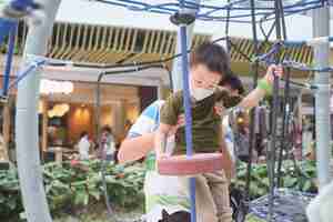 Photo cute little asian boy child wearing washable face mask climb on jungle gym at pulbic indoor playground with his father during covid-19 outbreak, new normal lifestyle concept - selective focus