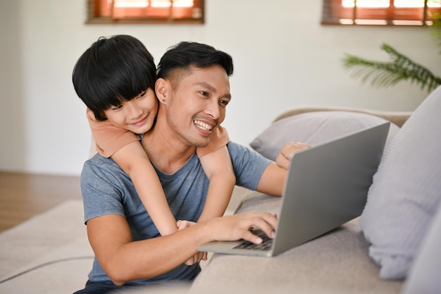 Photo cute little asian boy hugging his father from behind while he works on his laptop