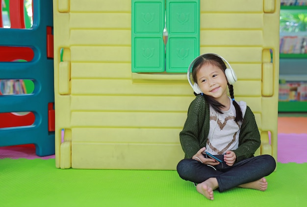 遊び場でおもちゃのプレイハウスの近くのヘッドフォンで音楽を聴くかわいい小さなアジア子供女の子