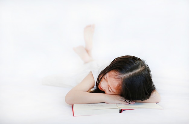 Милая маленькая азиатская девушка ребенка лежа на книге в твердом переплете на кровати над белой предпосылкой.