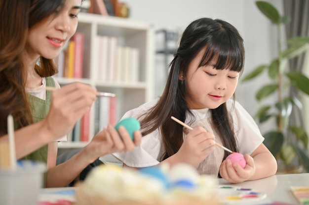 Cute little Asian girl painting Easter eggs with her mother in the living room