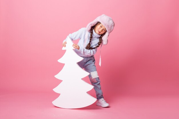 A cute little Asian girl in a winter hat stands next to a Christmas tree on a pink background. winter concept, space for text