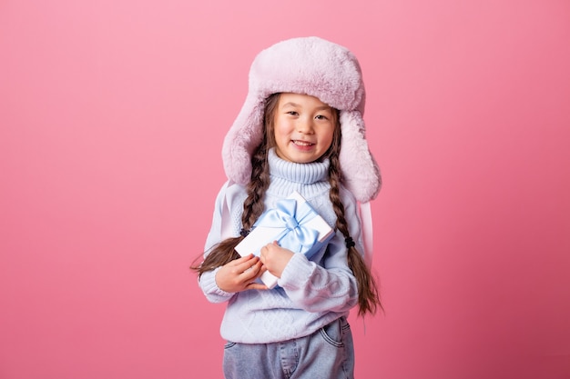 Cute little Asian girl in a winter hat and sweater holds a gift box. Christmas concept, text space