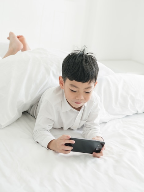 Il piccolo bambino asiatico sveglio ha messo a fuoco sullo smartphone mentre si trova sul letto