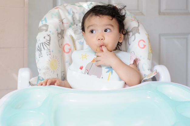 自分の指を吸って赤ちゃんの食卓に座っているかわいい小さなアジアの幼児の女の赤ちゃん。赤ちゃんの表現の概念