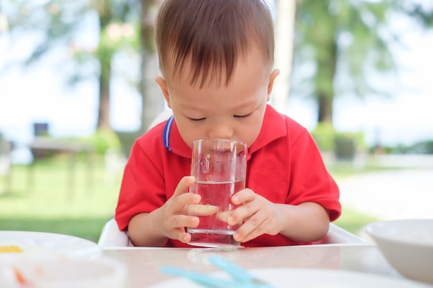 かわいいアジアの幼児男の子の子供がビーチリゾートのレストランで一人で水のガラスを保持＆飲むガラスに座って