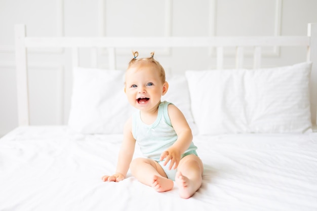 천연 면직물로 만든 바디수트를 입은 귀여운 아기가 침실에 있는 흰색 침대 린넨 위에 앉아 있습니다. 침대에서 행복한 작은 아기 어린이를 위한 직물 및 침대 린넨