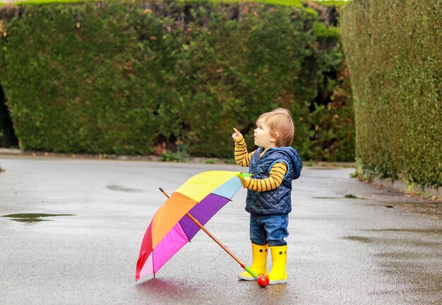 写真 濡れた路面でカラフルな虹の傘を持つ黄色のゴム製のブーツでかわいい小さな赤ちゃん