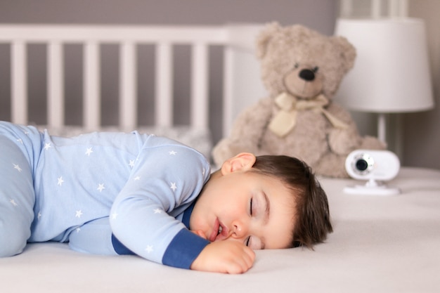 Cute little baby boy in light blue pajamas sleeping peacefully on bed with baby monitor 