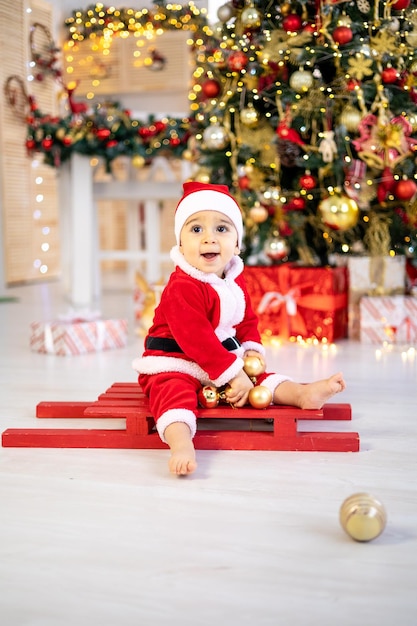 A cute little baby boy in a Santa costume is sitting on a sled under a festive Christmas tree with gifts in the living room of the house a happy kid is celebrating Christmas and New year at home