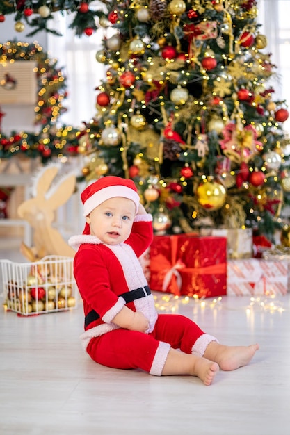 A cute little baby boy in a Santa costume is sitting with Christmas tree toys under a festive Christmas tree of the house a happy kid is celebrating Christmas and New year at home