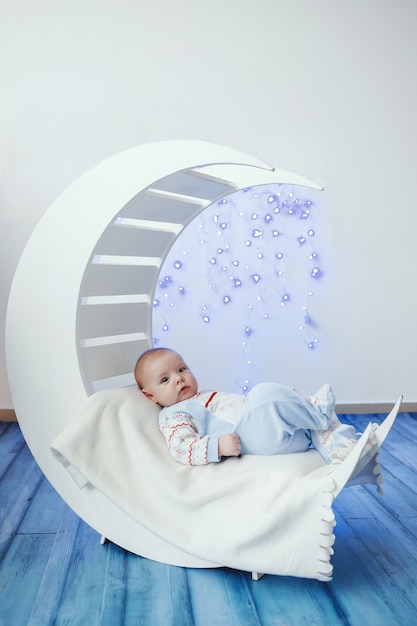 Piccolo neonato sveglio sul letto di legno bianco sotto forma di mezzaluna contro le luci di natale blu