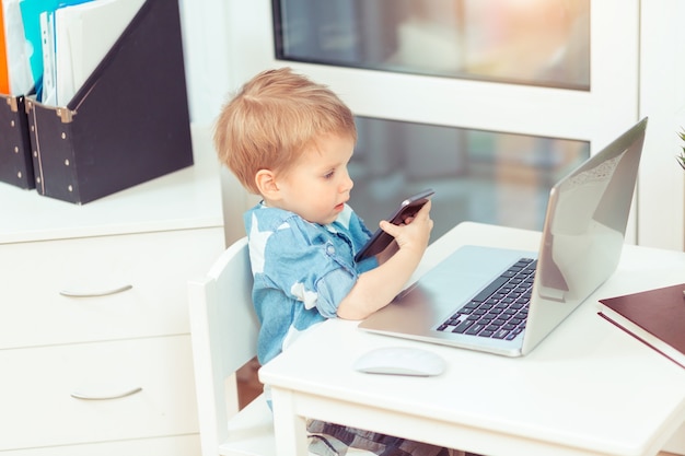 Cute little baby boy with computer laptop and mobile phone at home office Gadgets and kid concept