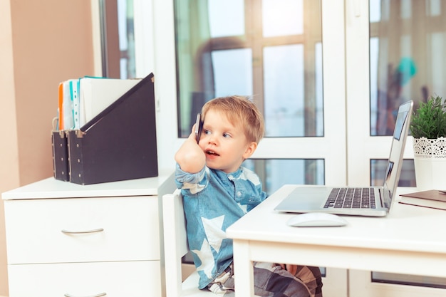 Cute little baby boy with computer laptop and mobile phone at home office smilin