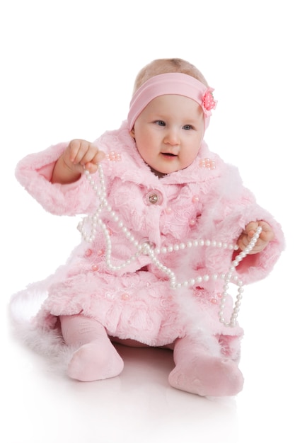 Photo cute little baby girl in pink jacket and  pants, sitting and playing with pearl beads isolated on the white background