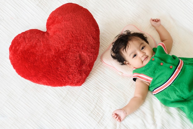 A cute little baby girl wear green dress lying beside a red heart shaped pillow on the bed